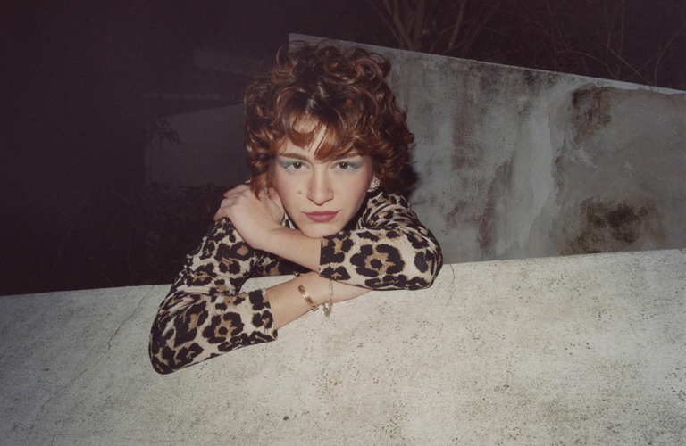 A woman wearing a leopard print dress leans over a wall, looking directly at the camera with a confident gaze. She has green mascara on her eyelashes, red curly hair and wears golden jewellery.