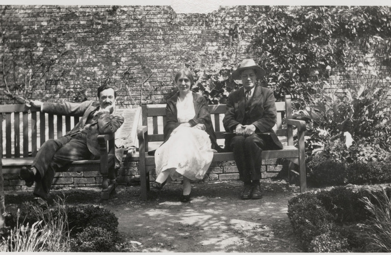 An old black and white photograph of three people (one woman, two men) sat on benches in walled garden, sunlight shining from above.