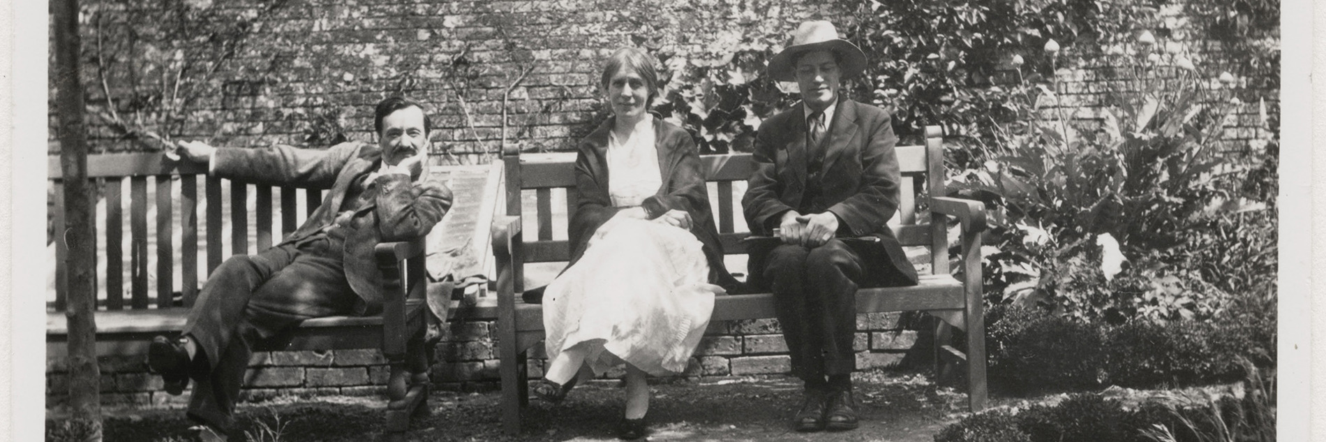 An old black and white photograph of three people (one woman, two men) sat on benches in walled garden, sunlight shining from above.