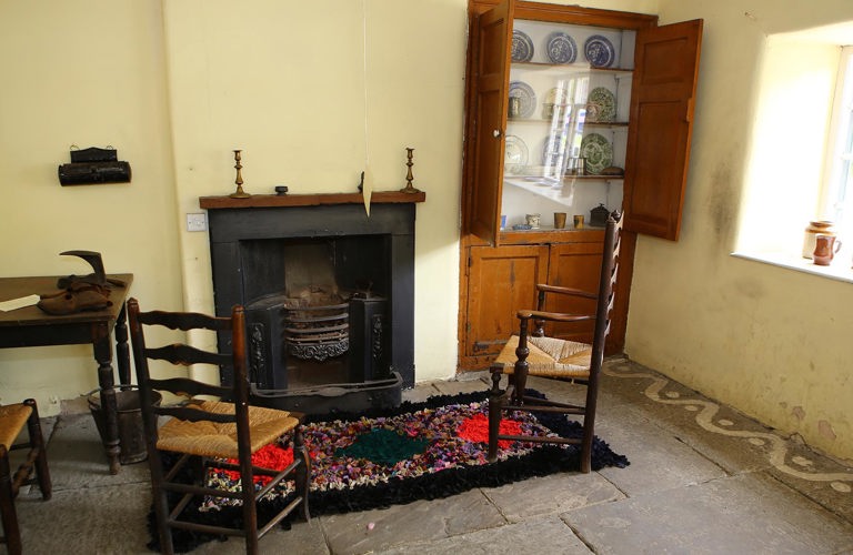 In a room with an open fire grate, two wooden ladderback chairs with woven rush seating stand by a pegged hearthside rug on a stone flagged floor. Alcove cupboards off to one side display antique china crockery. 