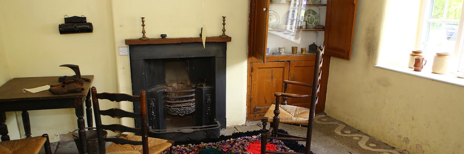 In a room with an open fire grate, two wooden ladderback chairs with woven rush seating stand by a pegged hearthside rug on a stone flagged floor. Alcove cupboards off to one side display antique china crockery. 