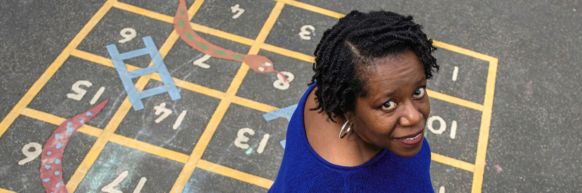 A photograph of a black woman wearing a knitted blue top, looking up at the camera directly into the lens. She is smiling and her silver hoop earring catches the light. The photo is taken from above, so we can see the woman is standing on a school playground game of snakes and ladders, painted directly onto the ground.