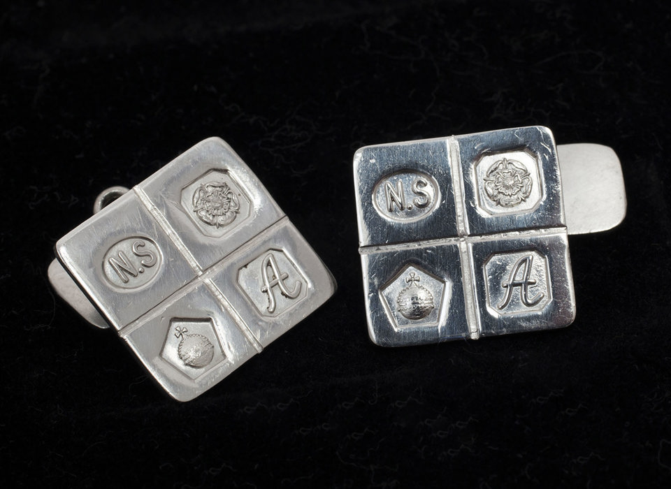 A pair of silver square cufflinks embossed with 4 symbols - the letters N.S, a flower, an orb and the letter A.  