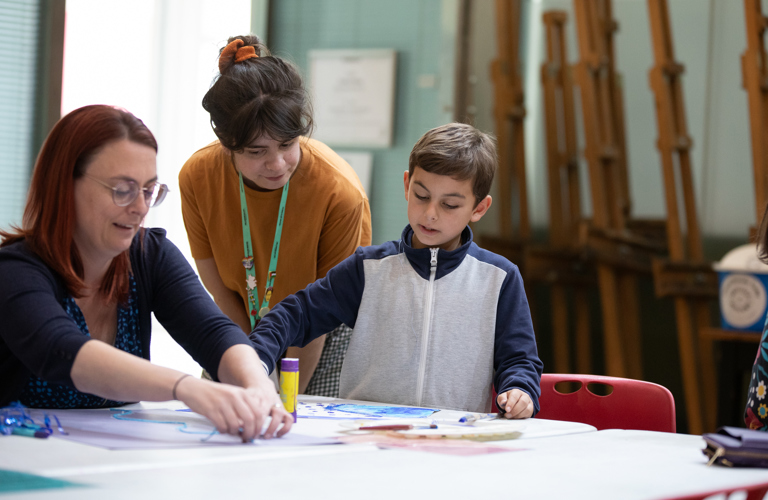 A photograph of a woman and a young boy crafting at a table. A woman with a Sheffield Museums lanyard is watching them.