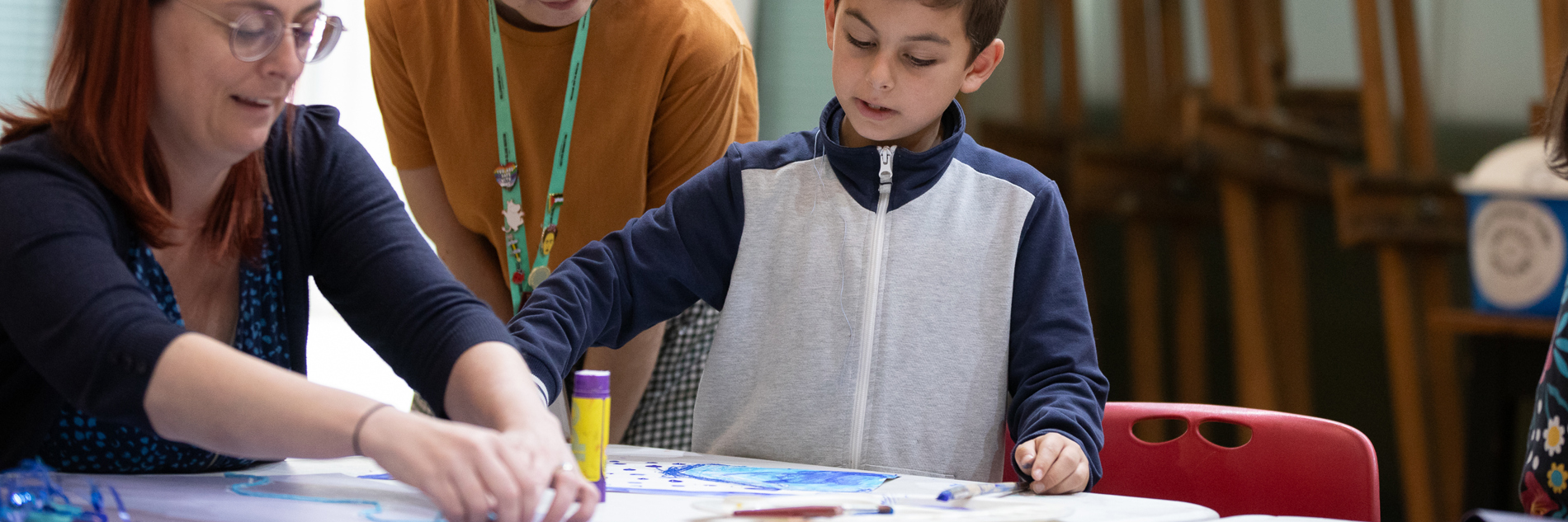 A photograph of a woman and a young boy crafting at a table. A woman with a Sheffield Museums lanyard is watching them.