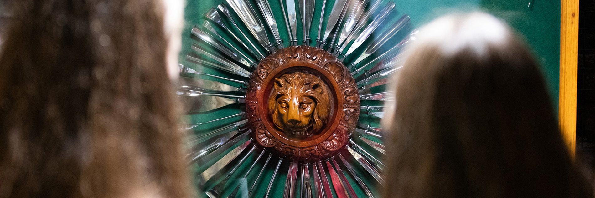 The back of two people's head as they look at a round wooden lion carving with metal tools arranged around it like sun rays.