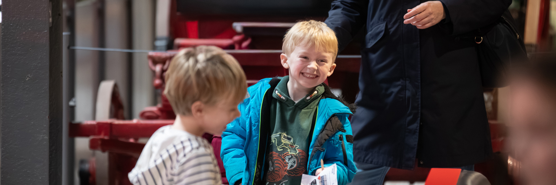 A photograph of two young boys smiling at Kelham Island Museum. The red frame of a carriage is visible behind them