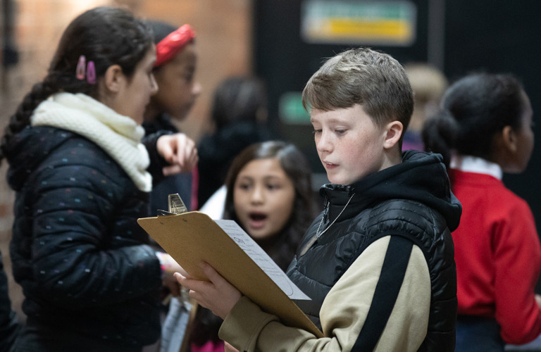 A group of primary school aged children, the child in focus is looking at a clipboard.