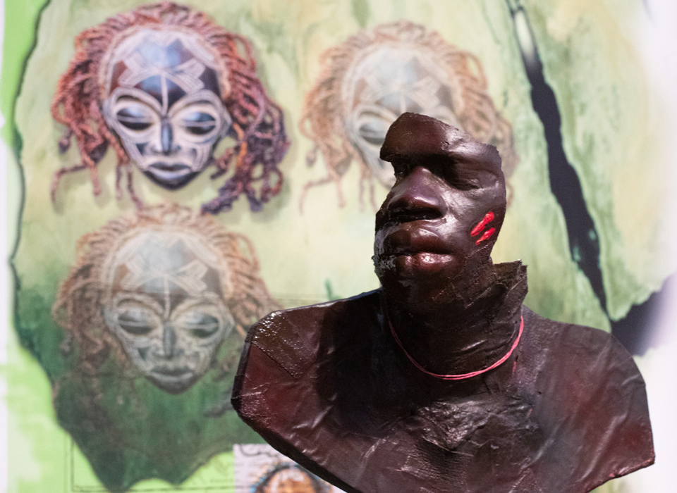 A detail photograph of Keith Piper's The Seven Rages Of Man. A broken bust of an black man is displayed in front of a graphic of tribal masks. 