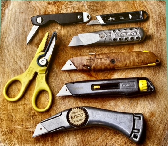 Two pen knives, 4 Stanley knives and a pair of scissors arranged on a table.