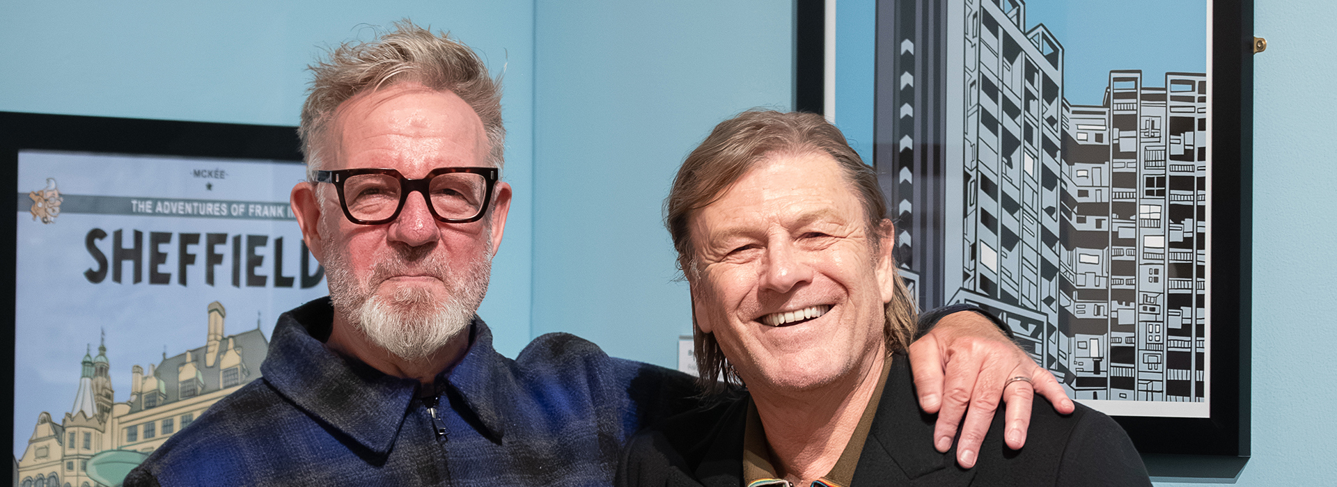 Two men stand arm in arm in front of paintings of Sheffield in an art gallery