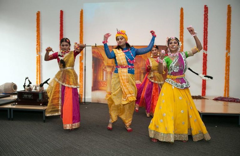 A photograph of three dancers wearing saris dancing. They all have their arms raised.