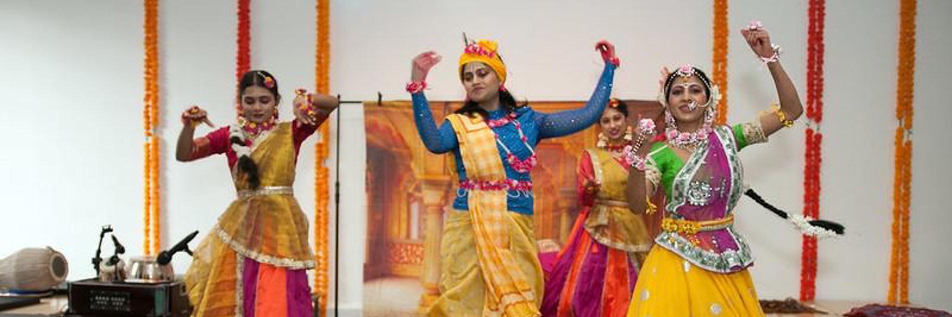 A photograph of three dancers wearing saris dancing. They all have their arms raised.