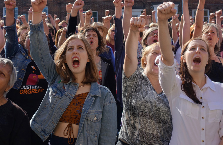 A photograph of a crowd of women at a protest or march. All have their mouths open, mid-shout. and have their right arms raised into a fist in the air. 