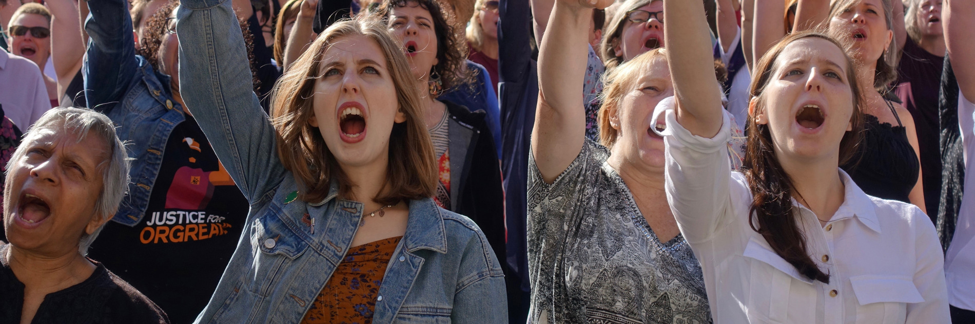 A photograph of a crowd of women at a protest or march. All have their mouths open, mid-shout. and have their right arms raised into a fist in the air. 