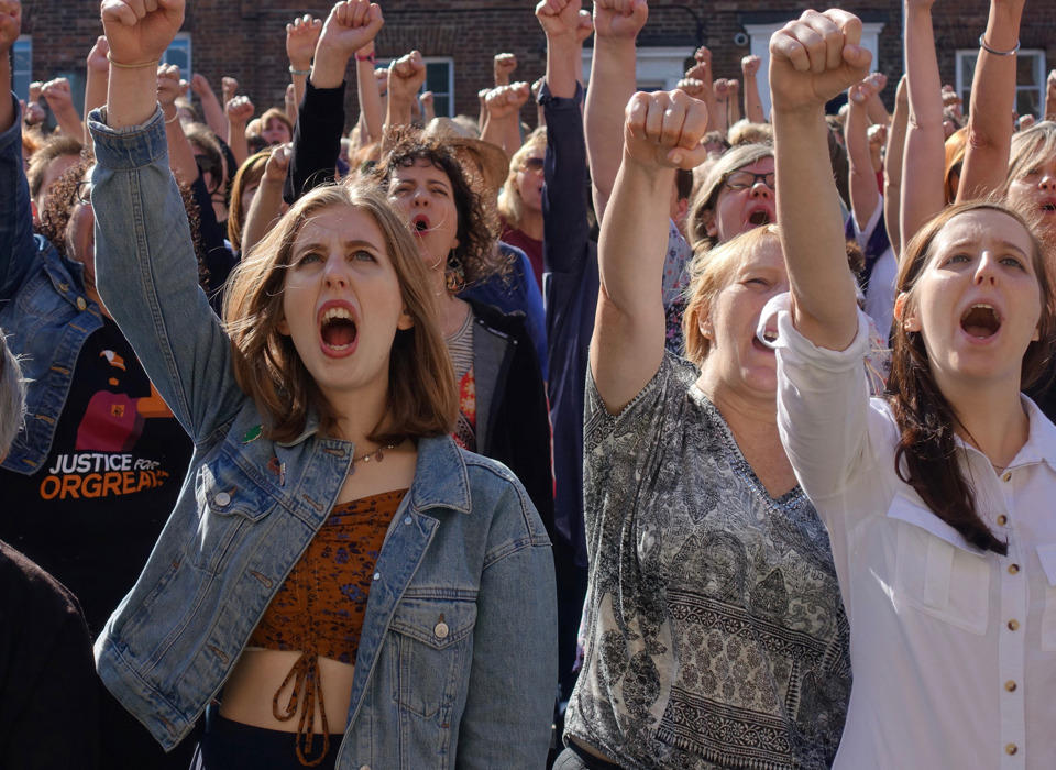 A photograph of a crowd of women at a protest or march. All have their mouths open, mid-shout. and have their right arms raised into a fist in the air. 