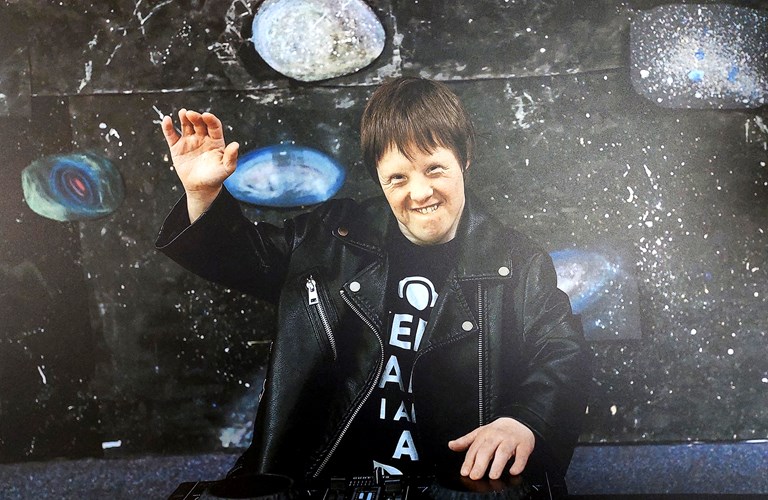 A smiling person stood behind DJ equipment waving. They stand in front of an abstract background that is reminiscent of space and stars.