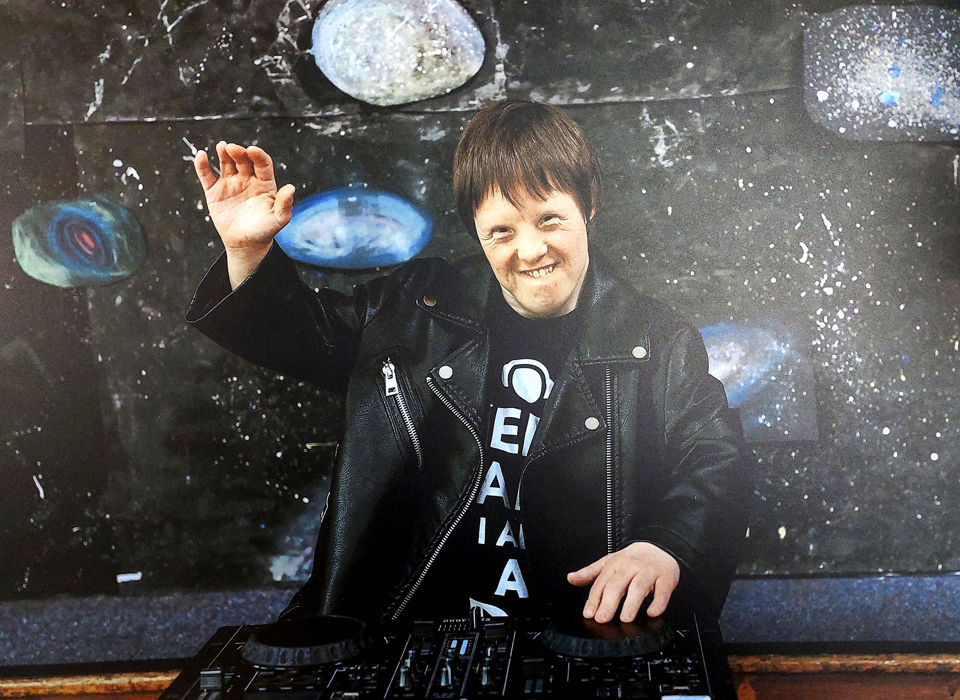 A smiling person stood behind DJ equipment waving. They stand in front of an abstract background that is reminiscent of space and stars.