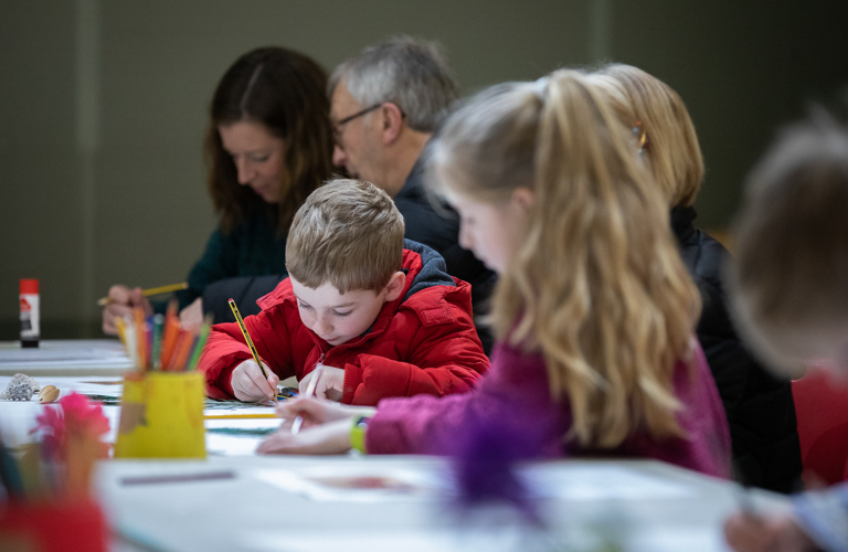 A photograph of a young boy drawing at a table. He is very focussed and there are other people sat at the table next to him, but they are out of focus. Paper and pots of pens are visible on the table in front of him