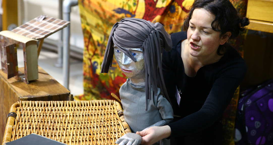 A person with a lifesized puppet, it is dressed in grey with grey fabric as hair, a small model of a building is on box behind them. 