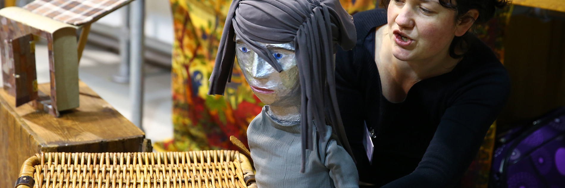 A person with a lifesized puppet, it is dressed in grey with grey fabric as hair, a small model of a building is on box behind them. 