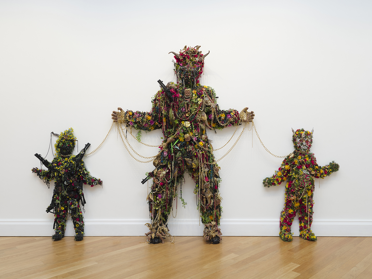 A photograph showing three of Hew Locke's Kingdom Of The Blind works, numbers 5, 6 and 7. Three humanoid shapes are shown, all covered in beads, chains and flowers. The central figure is large, adult size, and is linked through chains to two smaller, child size figures. The large figure, and the smaller black-coloured figure on the left both have large guns strapped to them. All three have horns coming out of their heads. They are all shown with their arms half raised. 