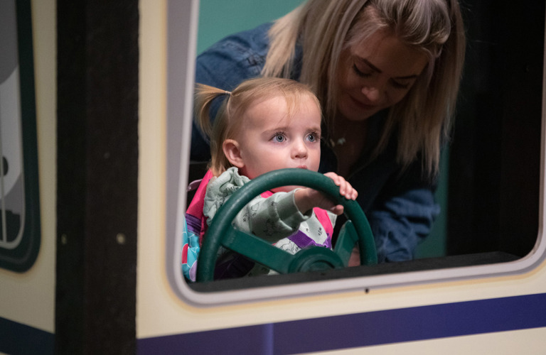 A young child and an adult sitting in a bus. The child is holding the bus' green steering wheel.