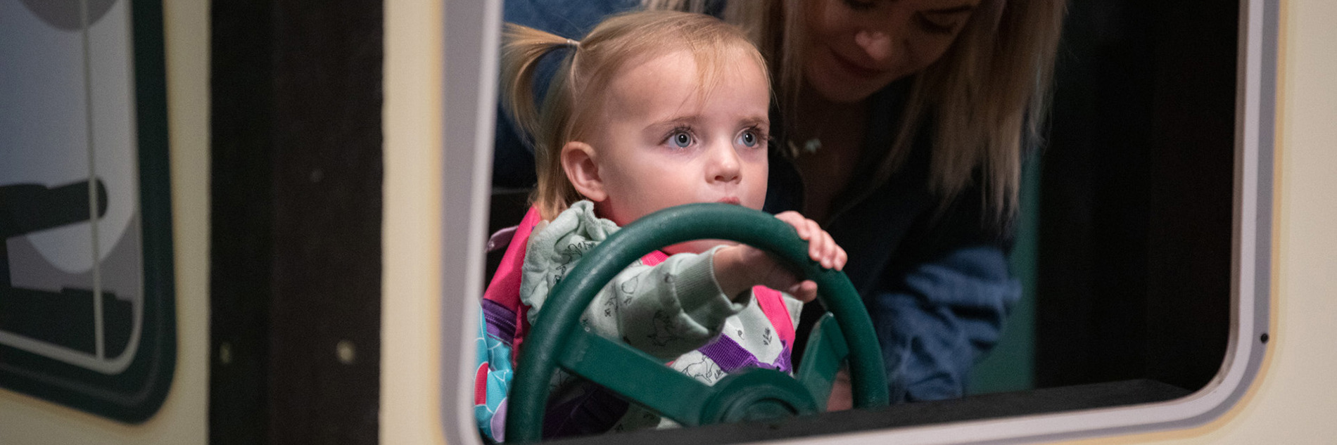 A young child and an adult sitting in a bus. The child is holding the bus' green steering wheel.