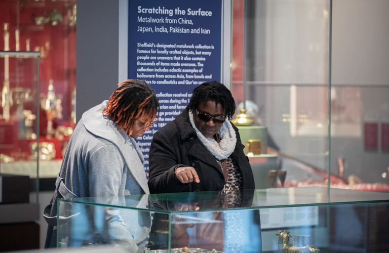 Two adults are looking at a glass cabinet in the gallery, one of the adults is pointing. There is a dark blue gallery sign behind them. 