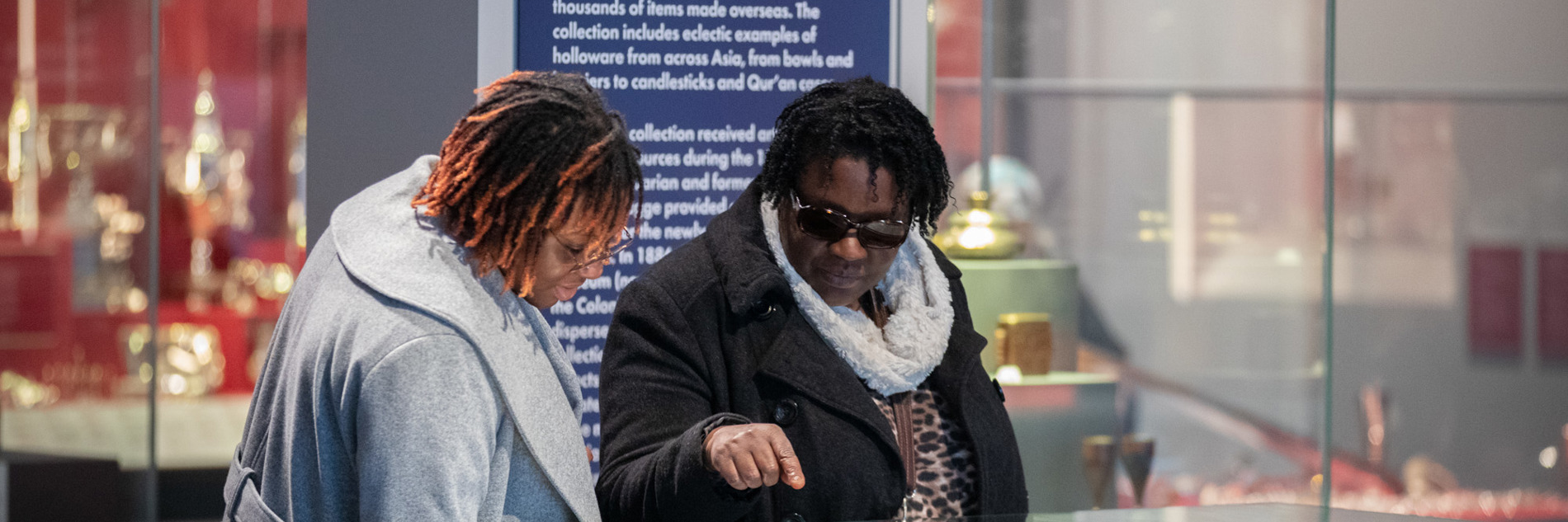 Two adults are looking at a glass cabinet in the gallery, one of the adults is pointing. There is a dark blue gallery sign behind them. 