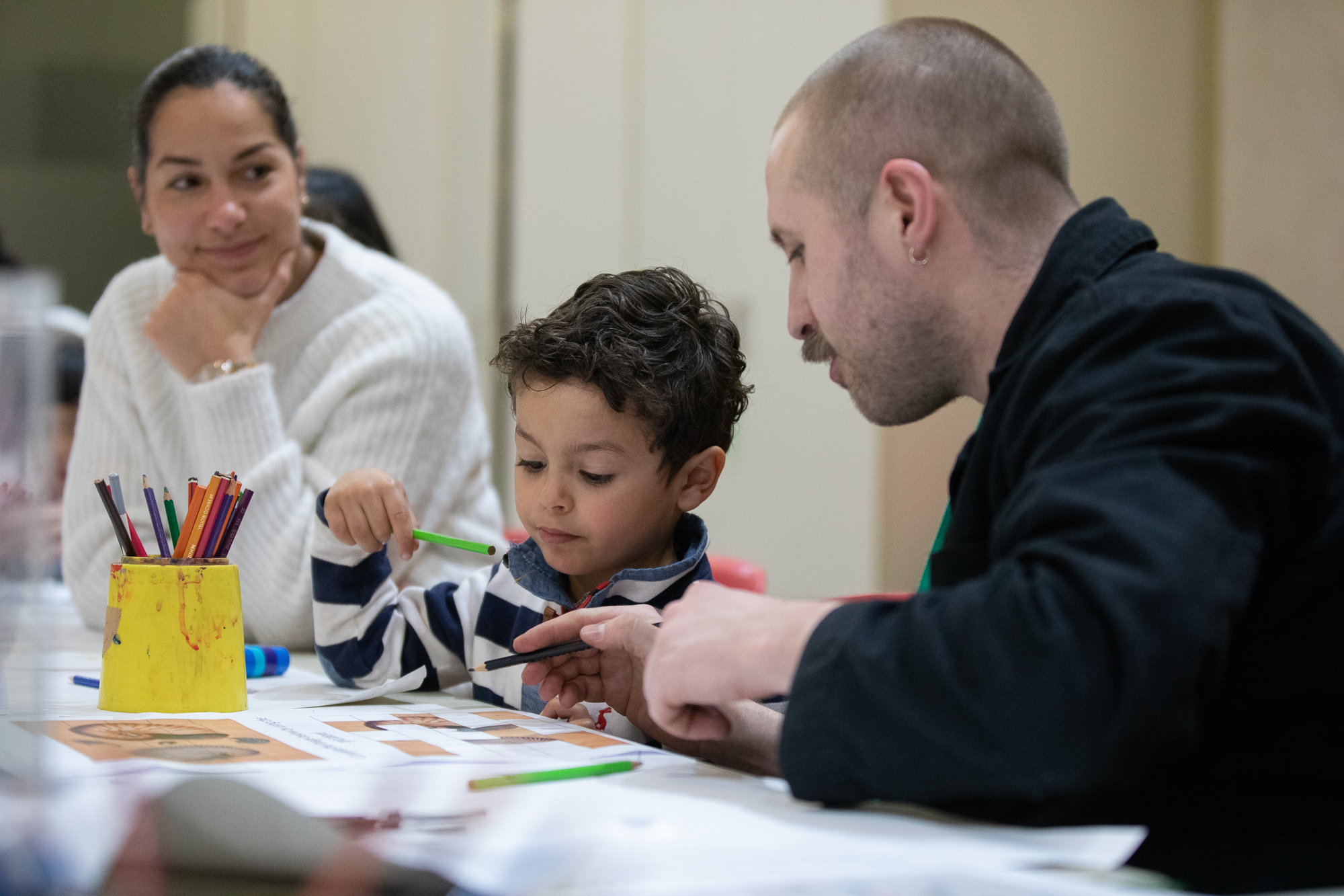 A photograph of a young boy sitting at a desk with a piece of paper in front of him and a green pencil in his hand. To the right, a woman watches on as a man instructs the child in an activity. A yellow pot of colourful pencils sits on the desk in front of them.