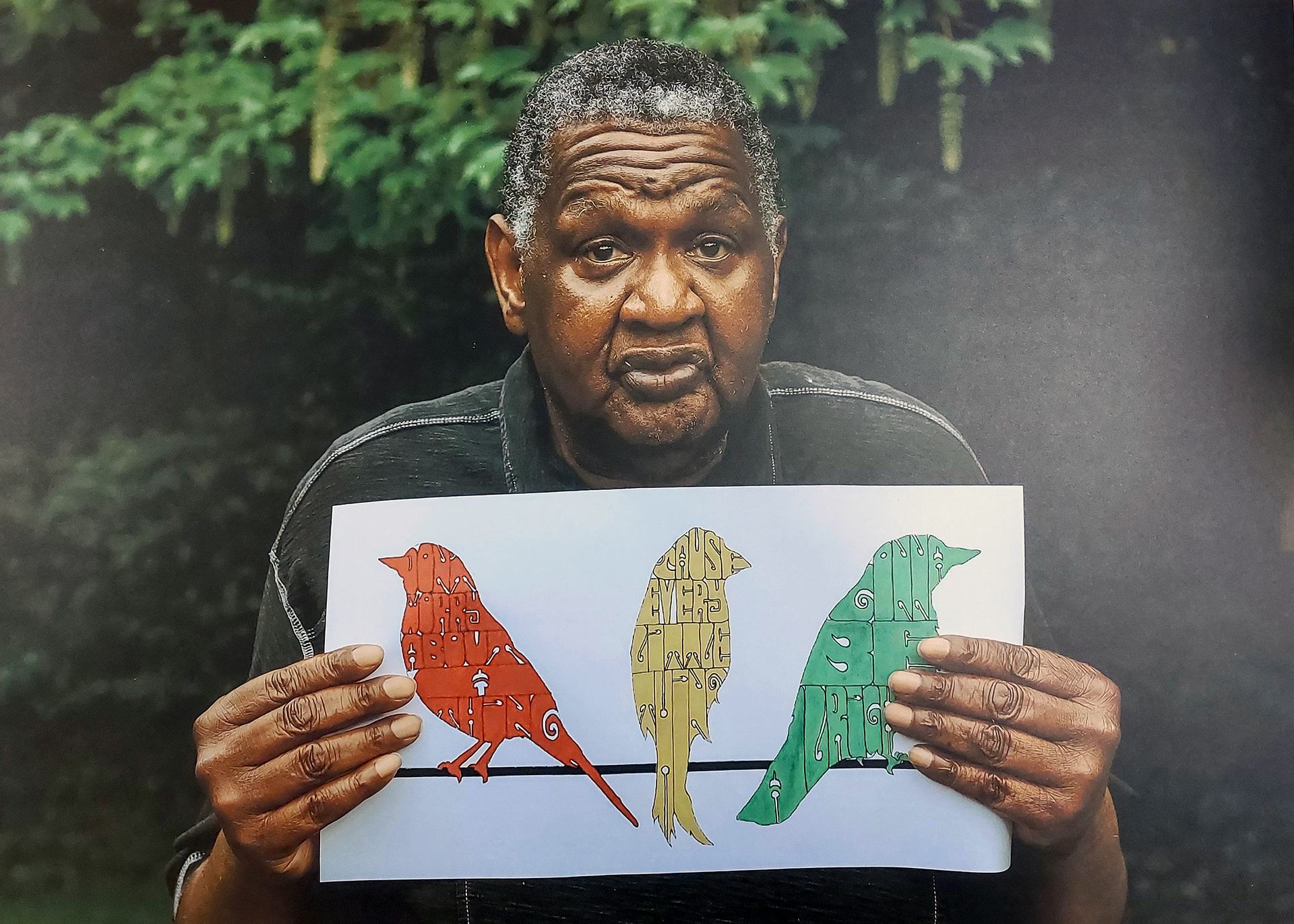 A photograph of a person holding up a drawing of three birds, one red, one yellow and one green. In the background of the photograph are trees and bushes,