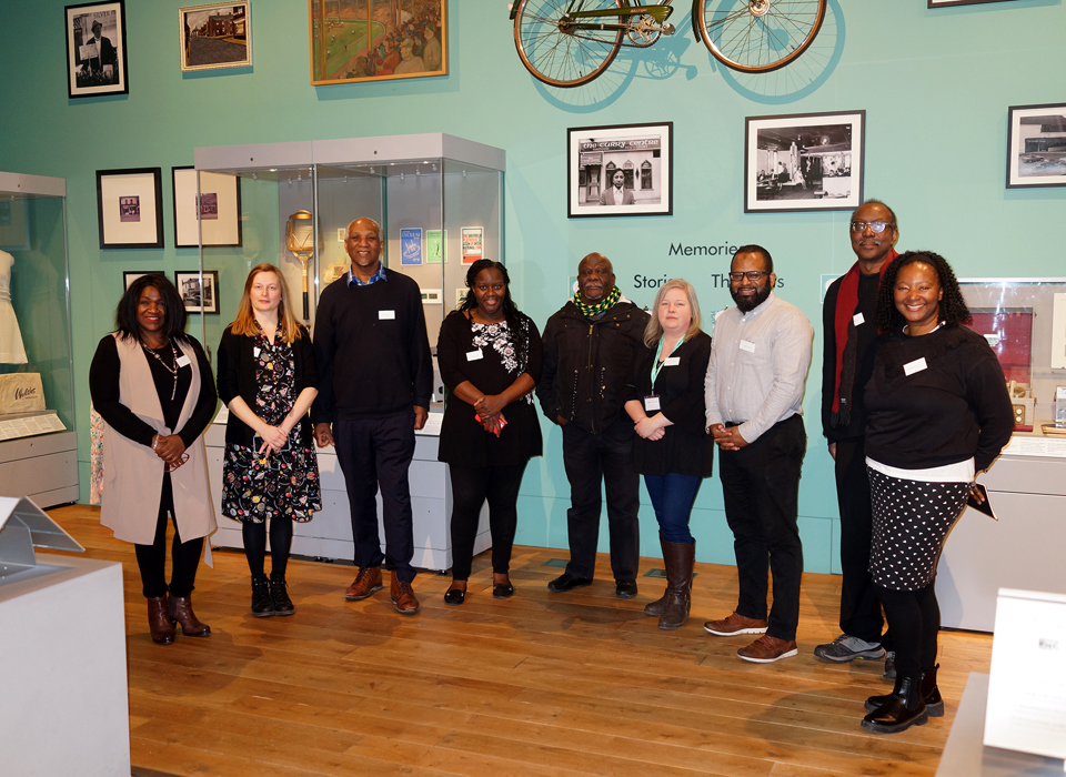 A group of nine people stood in a gallery. The floor is wooden and the gallery wall is painted eggshell blue. The group are surrounded by grey metal and glass display cases. on the wall behind them are several framed photographs and a bicycle suspended at height.