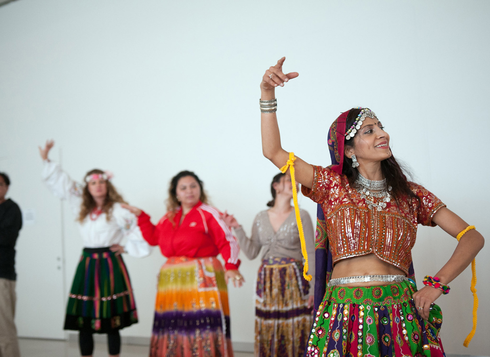 A photograph of five people dancing, with their right arms raised in the air, and their left placed on their hips. One person, probably the instructor, is shown at the front right of the image, while the others are slightly unfocused in the background. Four wear colourful skirts made out of a variety of fabrics.