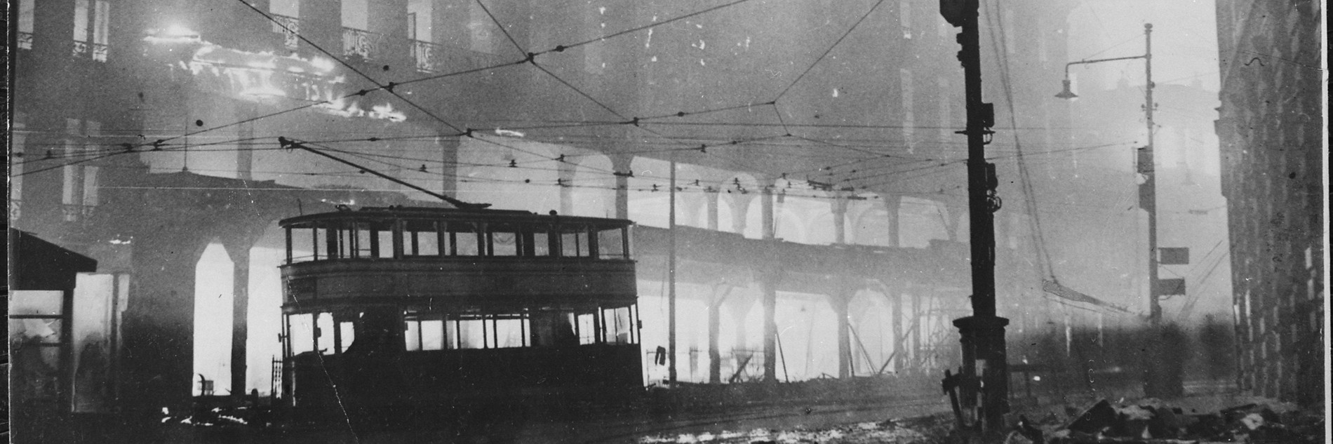 An old black and white photograph of Sheffield during the Blitz. A tram is shown off the track and there is rubble on the streets. The tram cables are visible and there is a large building behind.