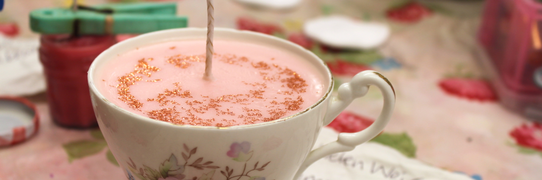 A photograph of a flowery teacup placed upon a flowery tablecloth. There is a pink candle set inside of it, with gold glitter on the surface and a fresh wick protruding from the centre. A handwritten papernote reveals the scent; lemongrass, lime and basil