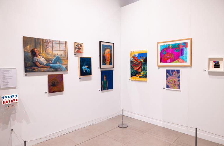 A photograph showing the corner of an exhibition. There are ten different portraits on the walls. A large one on the left of the image shows a woman relaxing in a sunlit window. Another shows a woman in a blue dress dramatically submerged in water. There is a rack of 3D glasses on the left of the wall, and barriers visible at the bottom of the walls.