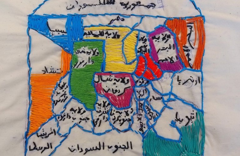 Embroidered outline of a house sectioned into brightly coloured shapes within and filled with words written in Arabic. 