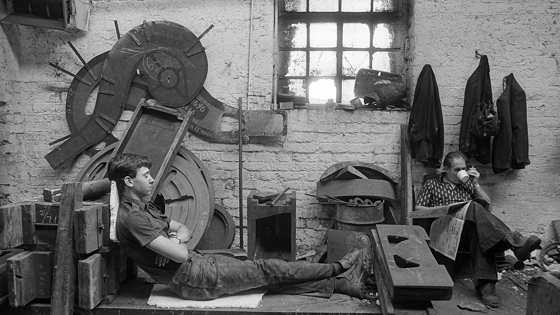 A black and white photograph of two adults in a workshop.  One adult is leaning against equipment with their eyes closed and the other adult is sitting in a chair, reading a newspaper and drinking out of a cup whilst holding a cigarette. 