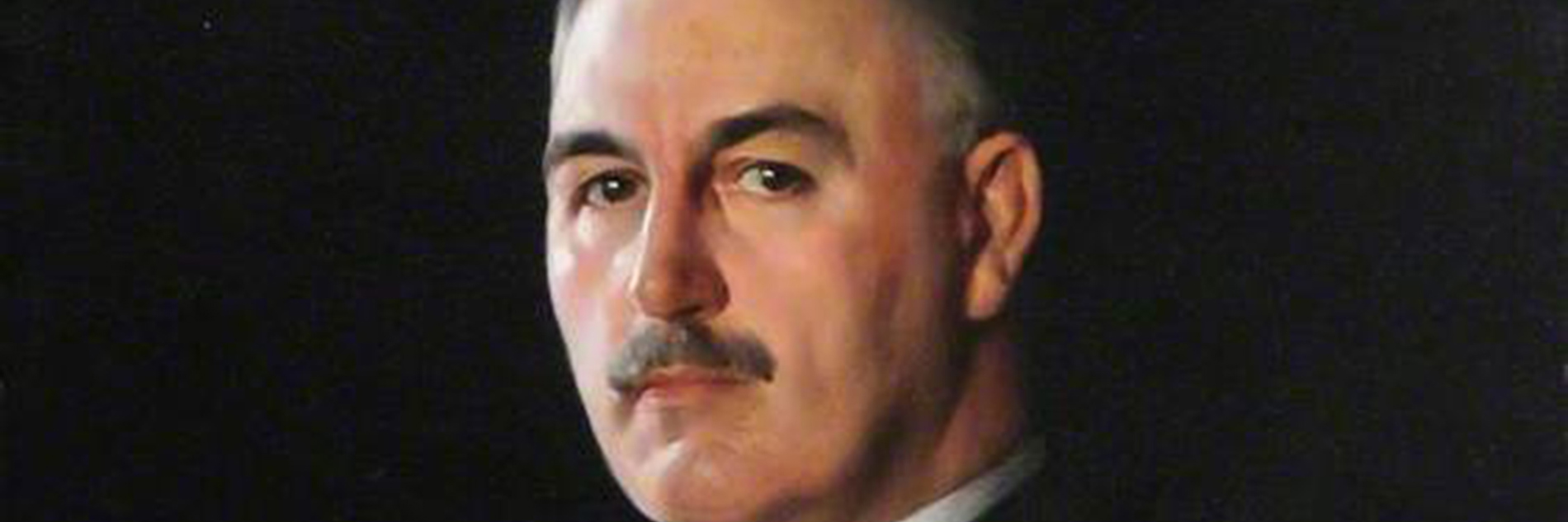 A portrait painting of Dr J.G Graves at around 77 years old. He looks out from the canvas directly with a serious expression and has short, grey hair, a neat moustache and he's wearing a dark suit againist a plain, black background. 