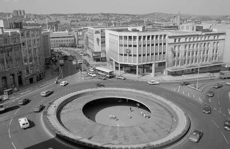 A black and white photograph of a roundabout (known as the “Hole in the Road”) with a view of Sheffield in the background. Cars and buses are driving round the roundabout, and in the middle, below street level, there are people walking and sitting.