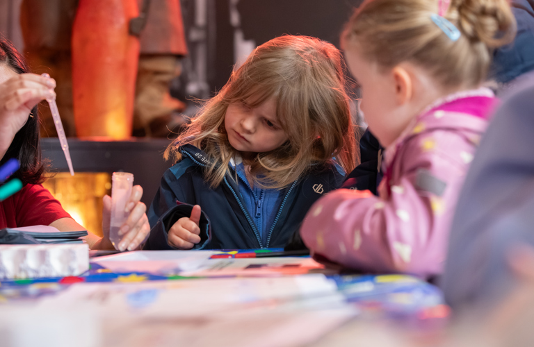 A photograph of two girls watching an adult do an experiment using a test tube and a pipette.