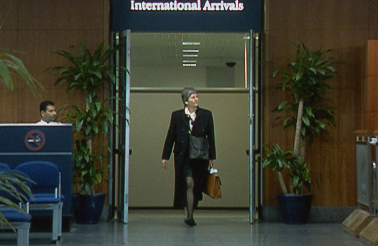 A video still of a smartly dressed person walking through a doorway marked "International Arrivals" at an airport terminal. To the left of the doorway there is a a person is sitting at a desk with a large "No Smoking Sign" on the front of it and chairs in front and to the right of the doorway a person is waiting behind a barrier.  