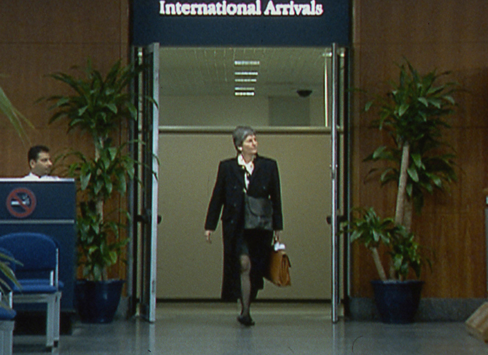 A video still of a smartly dressed person walking through a doorway marked "International Arrivals" at an airport terminal. To the left of the doorway there is a a person is sitting at a desk with a large "No Smoking Sign" on the front of it and chairs in front and to the right of the doorway a person is waiting behind a barrier.  