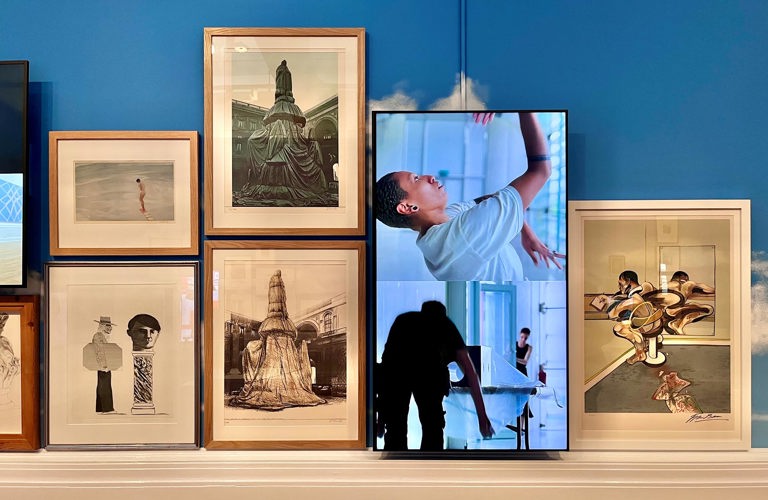 A blue wall with 5 framed artworks of differing sizes next to a screen showing a split screen - one screen shows an adult dancing and the other shows an adult moving an object whilst another adult holds a door open. 