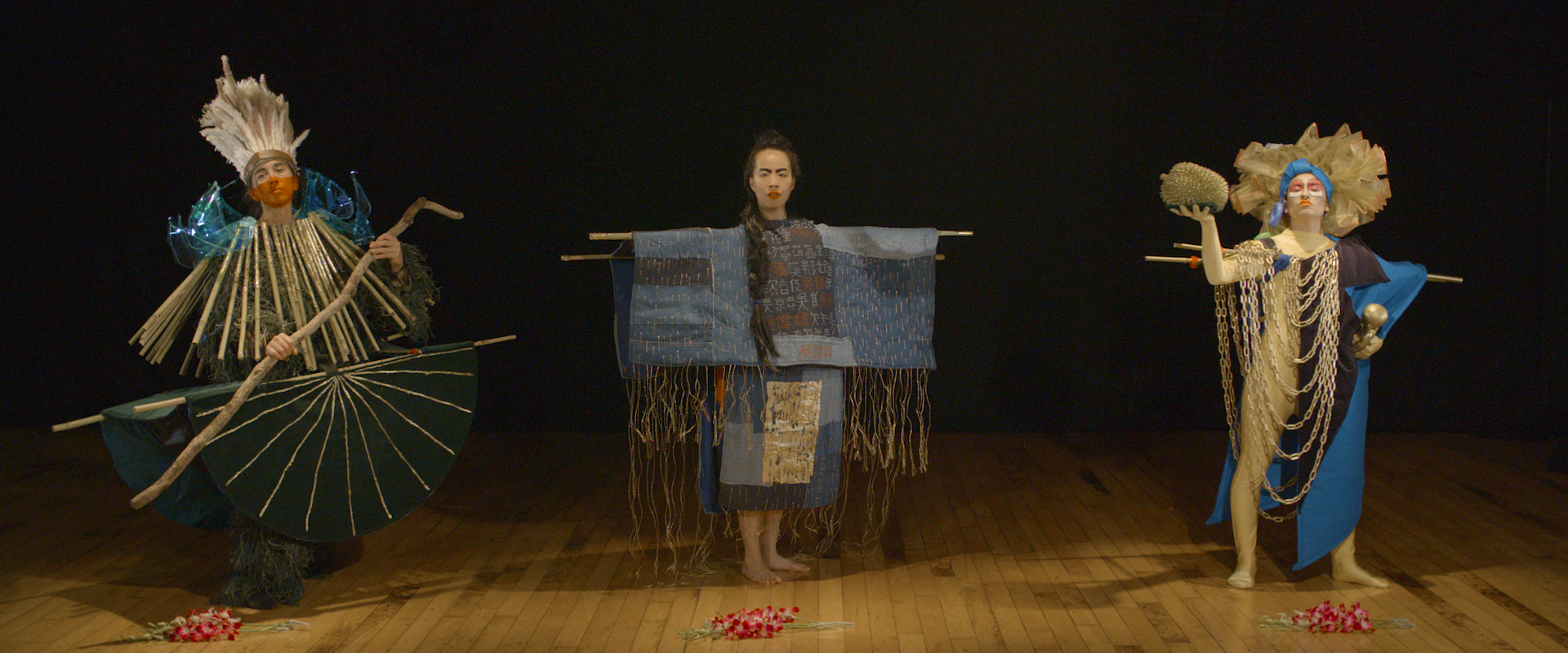 Three performers stand on a wooden floor, each wearing differently constructed costumes. Flowers are laid on the floor in front of them. 