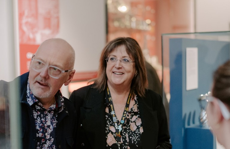 A photograph of two adults in a museum gallery. Other adults and object cases can be seen in the background. 