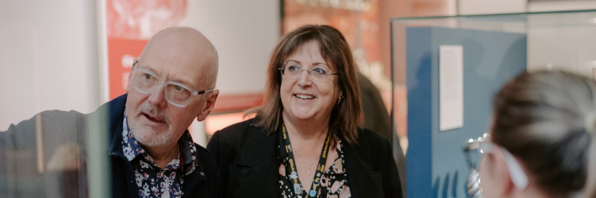 A photograph of two adults in a museum gallery. Other adults and object cases can be seen in the background. 