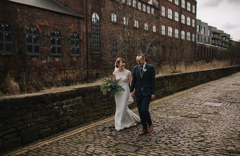 Two adults  in wedding outfits walking on the cobbled road outside Kelham Island Museum