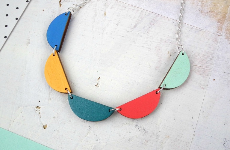 A photograph of a necklace consisting of a silver chain and five wooden scallop pieces in different colours. From left to right, the scallops are blue, yellow, turquoise, red and pale blue.